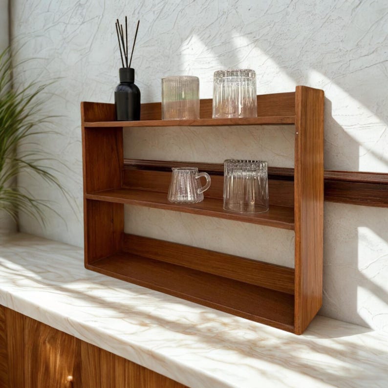Op de afbeelding: Een houten plank met drie niveaus en een warme bruine afwerking. De planken bevatten helder glaswerk, waaronder bekers en een kleine mok. Een zwarte fles met riet staat op de bovenste plank. De plank staat op een marmeren aanrecht.
