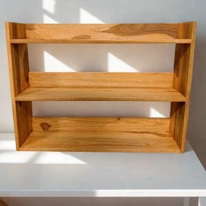 Op de afbeelding: Een houten plank met drie niveaus en een natuurlijke afwerking. De plank heeft een rechthoekige vorm met een licht schuin ontwerp aan de zijkanten. De planken zijn gelijkmatig verdeeld en bieden opbergruimte. De houtnerf is zichtbaar.