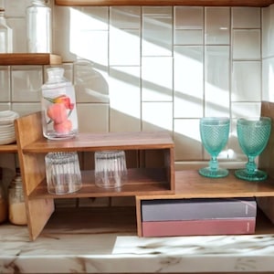 May include: A wooden tiered shelf unit with a clear glass jar containing pink fruit. Two clear drinking glasses and two turquoise goblets are displayed. The shelf sits on a marble countertop against a white tile backsplash.