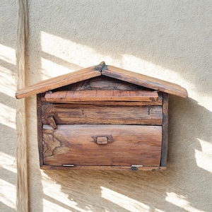 May include: A wooden mailbox with a pitched roof, mounted on a beige wall. The mailbox features a drawer with a small handle and a slot for letters. The wood has a natural, weathered appearance.