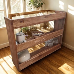 Op de afbeelding: Een houten plankeneenheid met drie niveaus, gemaakt van warm getint hout. De eenheid is gevuld met decoratieve items, waaronder een witte keramische container, een geweven mand en een witte vaas. De woorden "Back side" zijn zichtbaar op de plank.