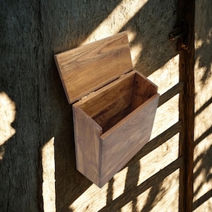 Puede incluir: Un buzón rectangular de madera con tapa con bisagras, montado sobre una superficie de madera texturizada. El buzón está abierto, revelando su interior. La madera tiene un tono cálido y natural, y la escena está iluminada por la luz del sol.