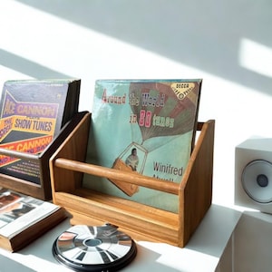 May include: A wooden vinyl record storage crate holding a record titled Around the World in 80 tunes by Winifred Atwell. The crate features a horizontal support bar and a natural wood grain finish, positioned next to another record holder and a speaker.