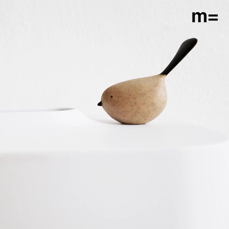 May include: A small, brown ceramic bird with a black tail sits on a white surface. The bird has a simple, hand-painted face.