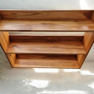 Op de afbeelding: Een houten plankeneenheid met drie niveaus en een natuurlijke houtnerf afwerking. De planken zijn rechthoekig en lijken te zweven, met een eenvoudig, modern ontwerp. De eenheid is waarschijnlijk voor opslag of weergave.