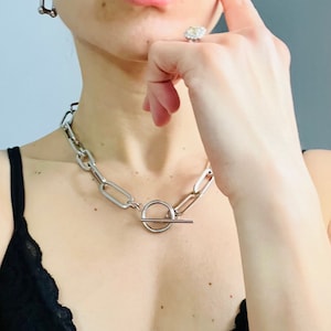 May include: A silver chain necklace with a toggle clasp. The necklace is thick and chunky, with large, oval links. The necklace is worn by a person with a black lace top.