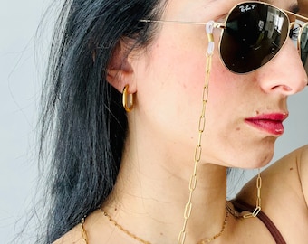 May include: A gold chain with a clasp on each end, worn as a glasses chain. The chain is hanging from the ears of a person wearing brown sunglasses and a brown tank top.