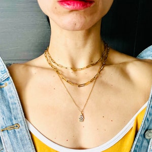 Può includere: Una collana a catena d'oro con tre strati di catene collegate e un ciondolo a forma di mano di Hamsa.