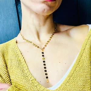 May include: A gold necklace with a long, dangling chain of small, round discs. The necklace is worn by a person with a yellow sweater.