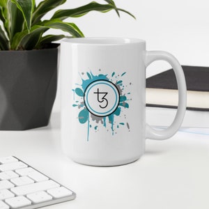 May include: White ceramic mug with a teal and gray paint splatter design. The center of the design features a circle with a gray outline and a black symbol of a cross with a number 3 inside.