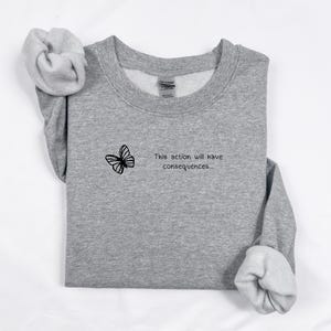 Puede incluir: Una sudadera gris jaspeado con un diseño de mariposa negra y el texto "This action will have consequences...". La sudadera tiene cuello redondo y mangas largas con puños enrollados.