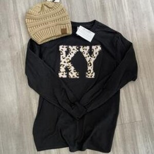 Peut inclure: Un t-shirt noir à manches longues avec les lettres "KY" dans un motif léopard. Un bonnet en tricot beige avec une petite étiquette est placé au-dessus du t-shirt. Les articles sont présentés sur une surface claire imitation bois.