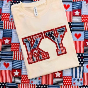 May include: A cream-colored short-sleeve t-shirt featuring the letters KY in a patchwork style. The letters are filled with various red, white, and blue patterns including stars and stripes, outlined in a thick red stitched border.