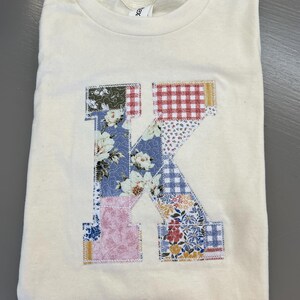 Pode incluir: Camiseta creme com uma grande letra "K" em patchwork aplicada na frente. A letra é composta por vários tecidos florais e estampados em tons de azul, rosa, vermelho e branco. A camiseta está cuidadosamente dobrada.