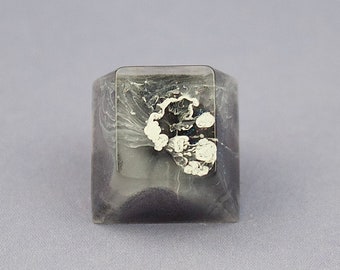 May include: A square-shaped ring with a clear resin base and a black and white design inside. The design looks like a cloud or a swirl.