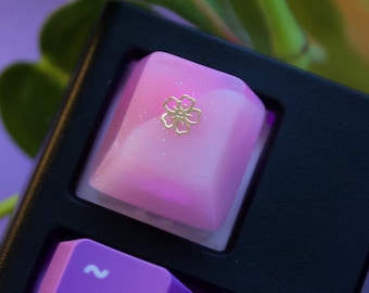 May include: Close-up of a mechanical keyboard with a custom keycap. The keycap is a gradient of pink and white with a gold floral design. Other keys are purple. The keyboard is black.