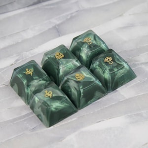 Könnte beinhalten: Sechs grüne und weiße marmorierte Keycaps mit goldenen Akzenten. Jede Keycap hat ein anderes Design, darunter eine Schlange, ein Pilz und ein Blatt.