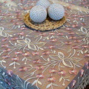 May include: A white lace tablecloth with a delicate floral embroidery pattern in pink and white. The tablecloth has a scalloped edge and is decorated with three white decorative balls.