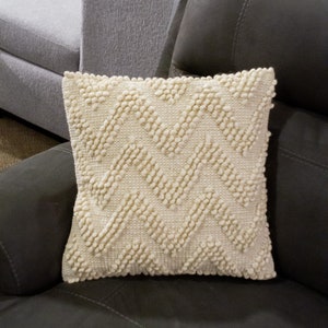 May include: A cream-coloured throw pillow with a textured diamond pattern. The pillow is made of a soft, fluffy material and is perfect for adding a touch of comfort and style to any room.