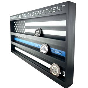 Puede incluir: Un expositor negro con forma de bandera estadounidense, con las palabras "LOS ANGELES POLICE DEPARTMENT" en la parte superior. Cuenta con una franja azul y contiene varias monedas. El texto "OFFICER" y "#12345" son visibles.