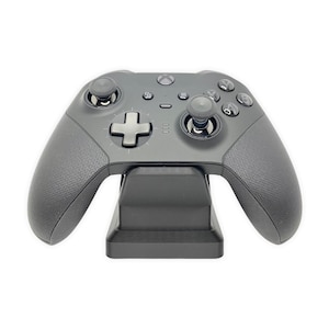 Może przedstawiać: Czarny kontroler Xbox znajduje się na czarnym stojaku ładującym. Kontroler ma teksturę i czarną i srebrną płytę czołową. Stojak ma prostokątną podstawę i podwyższoną platformę dla kontrolera.