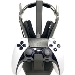 Peut inclure: Un support de charge pour manette PlayStation 5 noir et blanc avec un support de casque noir. La manette est blanche avec des accents noirs et le casque est noir avec des accents argentés.