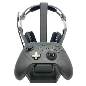 Könnte beinhalten: Ein schwarzer Gaming-Headset-Ständer mit einem schwarzen Controller. Der Ständer hat eine gebogene Form, um das Headset zu halten, und eine Basis, um den Controller zu halten.