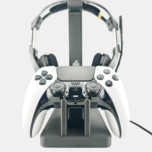 May include: A white and black gaming controller and headset set on a gray stand. The controller has black buttons and joysticks. The headset is black and silver. The stand is gray and has a charging port.