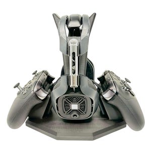 Może przedstawiać: Czarny stojak na słuchawki do gier z dwoma czarnymi kontrolerami po obu stronach. Stojak ma sześciokątną podstawę i czarno-szary wzór.