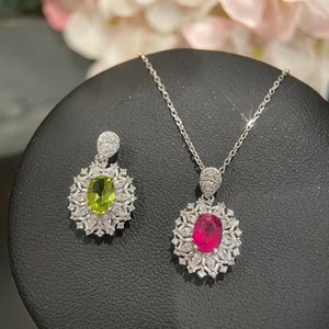 Puede incluir: Un conjunto de collar y pendientes de plata con un colgante de piedra roja y un colgante de piedra verde. Ambos colgantes están rodeados de piedras pequeñas transparentes.