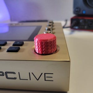 May include: A close-up of a red knob with a diamond pattern on a gold music production device. The device has the text "PC LIVE" printed on the front.