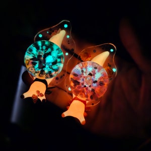 May include: Two glowing resin snail charms with a translucent shell and a detailed, colorful design. The snail on the left has a teal and black shell, while the snail on the right has a pink and orange shell.