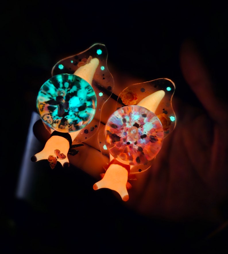 May include: Two glowing resin snail charms with a translucent shell and a detailed, colorful design. The snail on the left has a teal and black shell, while the snail on the right has a pink and orange shell.