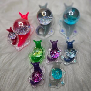 May include: A set of eight colorful snail figurines with iridescent glitter accents. Each snail has a clear dome on its back, filled with glitter and a variety of colors. The snails are arranged on a white fluffy surface.