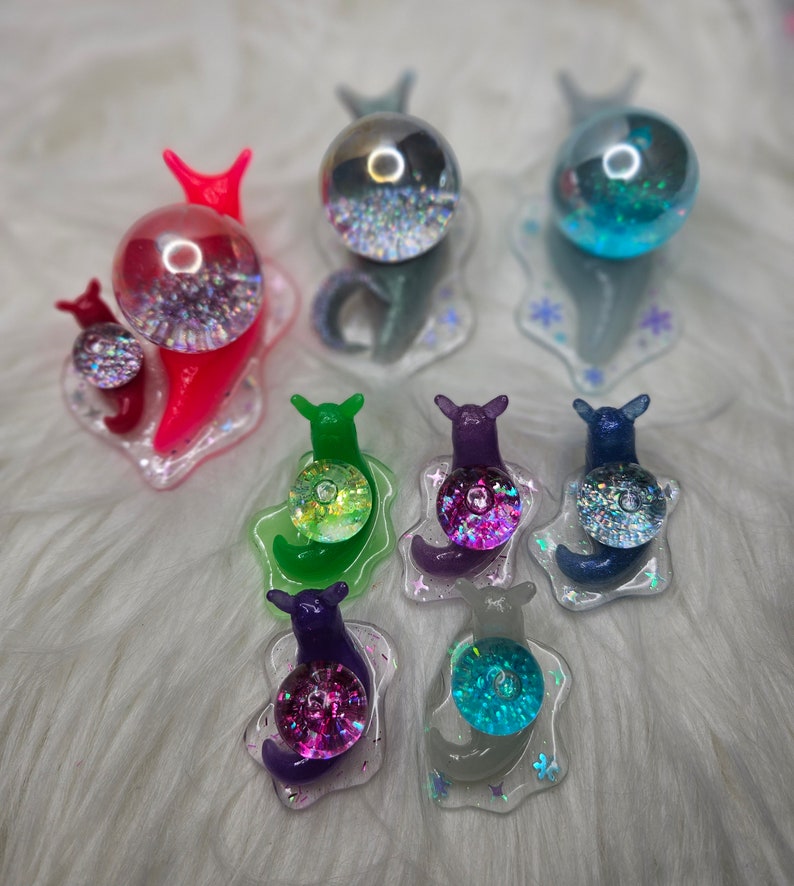 May include: A set of eight colorful snail figurines with iridescent glitter accents. Each snail has a clear dome on its back, filled with glitter and a variety of colors. The snails are arranged on a white fluffy surface.