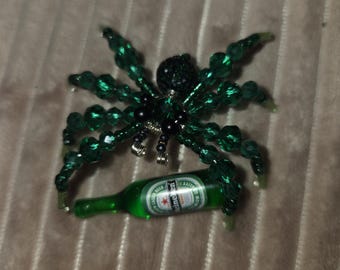 May include: A spider-shaped ornament crafted from green faceted beads and black beads, featuring a miniature green glass Heineken beer bottle held within its legs. The spider has a textured black head and silver wire accents.
