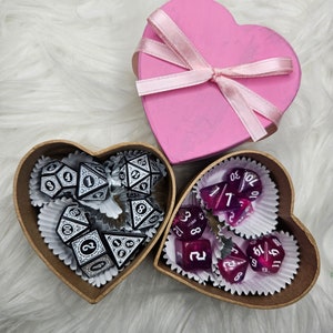 May include: A pink heart-shaped box tied with a light pink ribbon sits above two heart-shaped wooden boxes. One box contains black and white dice, the other contains purple dice. The dice are for tabletop games.