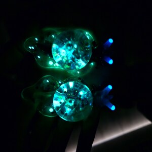 May include: Two clear glass ornaments with a blue and green iridescent glitter inside. The ornaments are shaped like fish with a tail fin and a small fin on top.