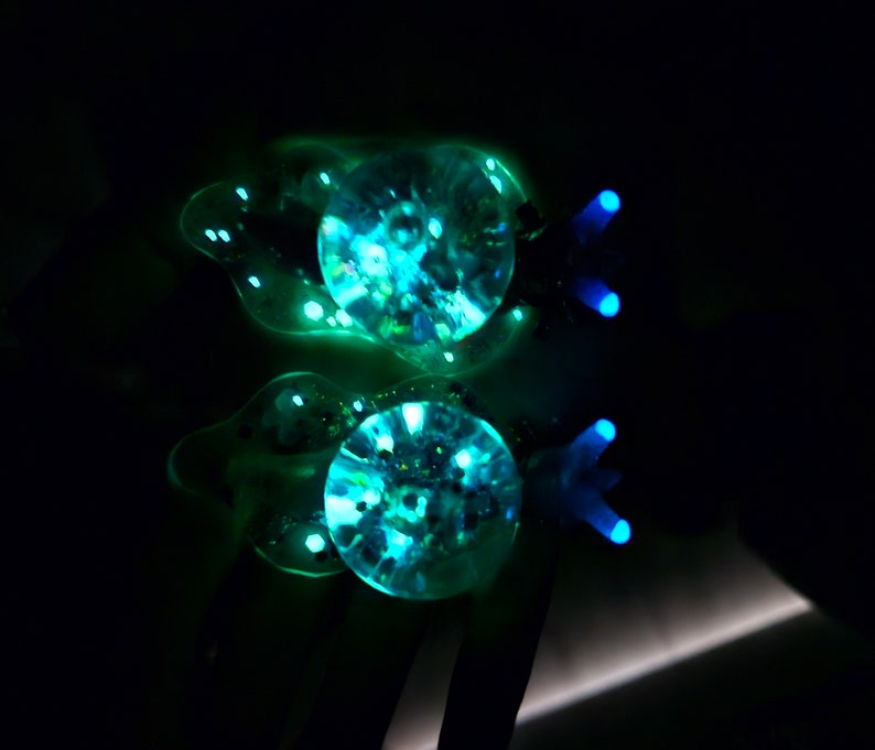 May include: Two clear glass ornaments with a blue and green iridescent glitter inside. The ornaments are shaped like fish with a tail fin and a small fin on top.