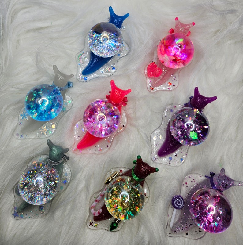 May include: A collection of ten colorful snail charms with glitter and iridescent details. Each snail charm has a different color and design, including pink, blue, purple, and green.