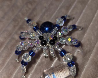 May include: A beaded spider figurine crafted with iridescent clear and deep blue beads, featuring a dark blue spherical body and two black eyes. The spider is positioned next to a miniature clear glass bottle labeled Absolut Vodka.