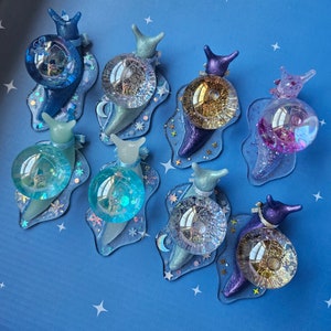 May include: A set of nine iridescent snail charms with different colored shells and glitter accents. Each snail charm has a clear glass bead on its back.