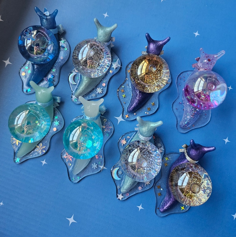 May include: A set of nine iridescent snail charms with different colored shells and glitter accents. Each snail charm has a clear glass bead on its back.