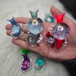 May include: Five colorful snail charms with iridescent glitter inside. The snails are made of resin and have a variety of colors, including blue, green, red, purple, and turquoise.