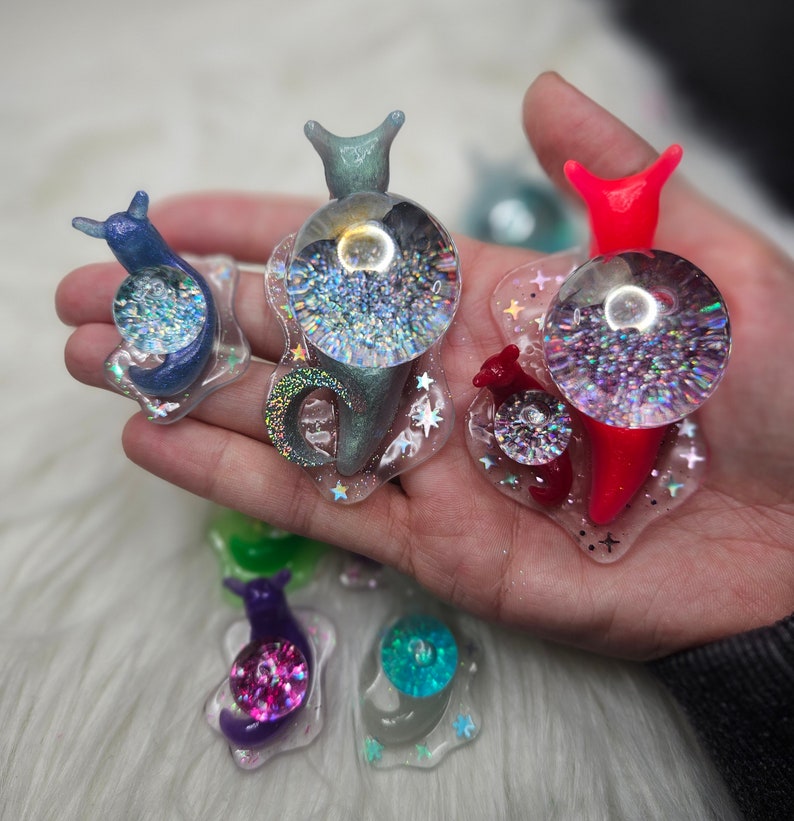 May include: Five colorful snail charms with iridescent glitter inside. The snails are made of resin and have a variety of colors, including blue, green, red, purple, and turquoise.