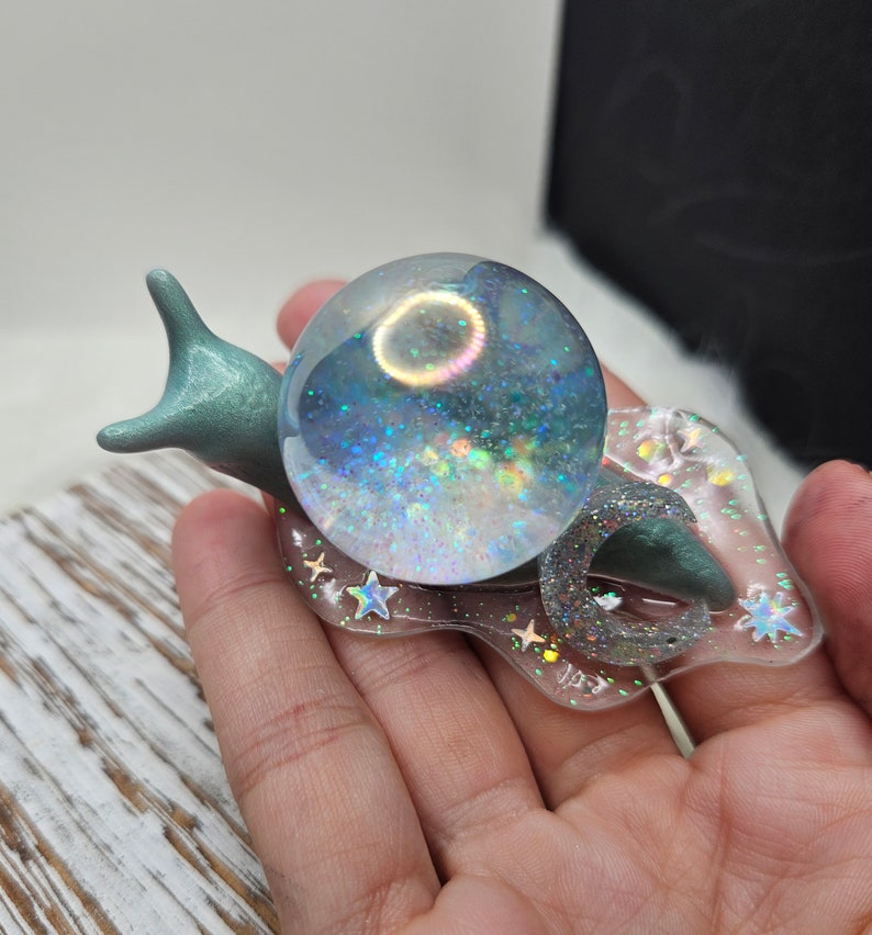 May include: A teal blue snail figurine with a clear iridescent glitter sphere on its back. The sphere is filled with iridescent glitter and stars. The snail is sitting on a clear resin base with a glittery finish.