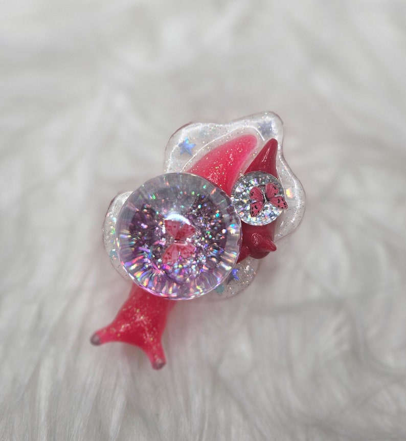 May include: A pink and white resin snail charm with a clear, glittery dome and a pink butterfly accent. The snail has a detailed shell and a whimsical design.