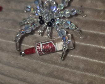 May include: A spider-shaped ornament crafted from iridescent clear beads with two black bead eyes. The spider holds a miniature clear glass bottle with a red cap and a label that reads Smirnoff Vodka in its front legs.