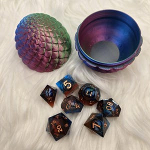May include: A dragon egg-shaped dice box in shades of blue, green, and purple, with a matching interior. Seven multi-sided dice with blue and brown accents are scattered nearby. The dice have numbers on them.