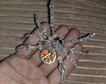 May include: A spider ornament crafted from clear, iridescent faceted beads. The body features a large, round, light-reflecting crystal, while the eight legs are constructed from smaller beads. Two small black beads serve as the eyes.